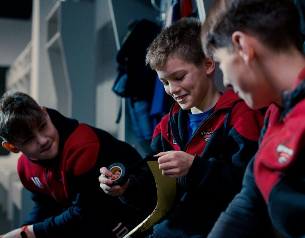 Three young hockey players in a dressing room, preparing equipment for a game.